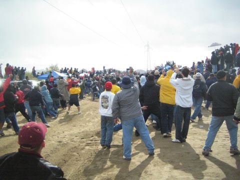 1990 Ford Bronco - large crowd of spectators spreads up a sandy hillside, raising cameras to watch race action under a power transmission tower and overcast s…