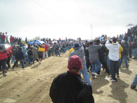 1990 Ford Bronco - spectator in a maroon cap looks out over a large crowd gathered along a sandy desert course under an overcast sky.