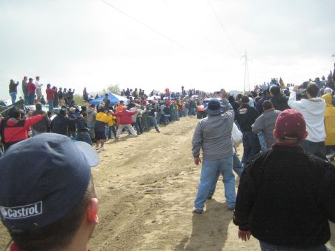 1990 Ford Bronco - Bundled-up spectators, one in a Castrol cap, stand along the edge of a sandy course watching distant crowds and tents.