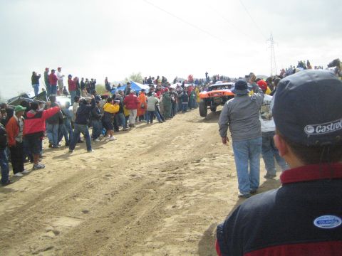1990 Ford Bronco - orange off-road race truck drives along a sandy desert course as a line of spectators watches from the left.