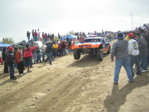 1990 Ford Bronco - orange trophy-style off-road truck crests a sandy berm flanked by crowds of spectators under a gray sky.