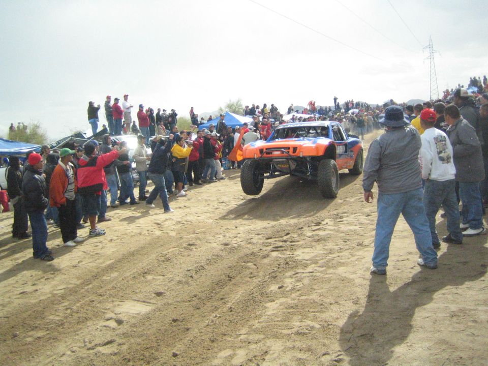 Orange off-road truck cresting a sandy rise with crowds on both sides