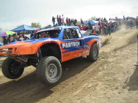 1990 Ford Bronco - orange and blue off-road trophy truck marked Fabtech, Fox and Toyo Tires kicks up dust on a sandy course before spectators.
