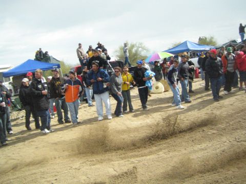 1990 Ford Bronco - crowd of bundled spectators stands along a sandy track beside blue and purple pop-up canopies on an overcast day.