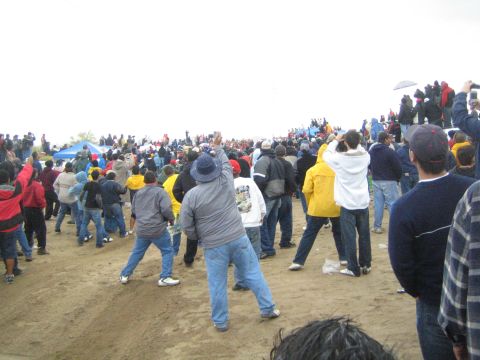 1990 Ford Bronco - large crowd of spectators cheers with raised arms along a sandy desert rise beneath a pale overcast sky.