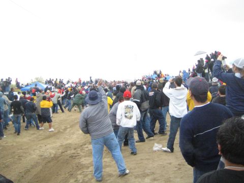 1990 Ford Bronco - Spectators in winter jackets and caps watch and photograph activity across a broad sandy field under gray skies.
