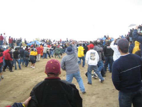 1990 Ford Bronco - spectator in a maroon cap stands amid a large crowd watching across a sandy desert course on a cloudy day.