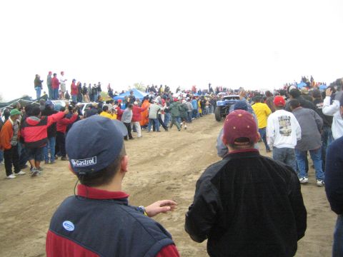 1990 Ford Bronco - Two spectators in caps watch from the foreground as a distant off-road truck travels a sandy course lined with crowds.