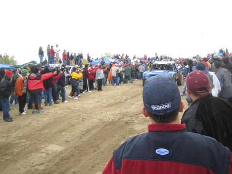 1990 Ford Bronco - blue off-road race truck approaches along a sandy course as spectators in caps watch from the foreground.