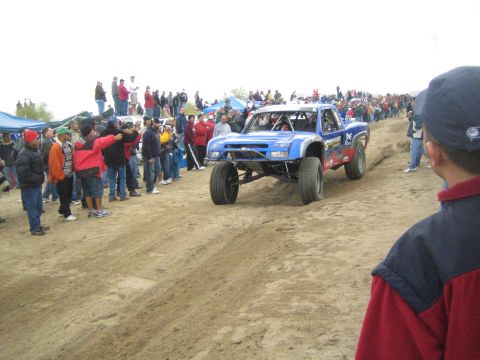 1990 Ford Bronco - blue off-road race truck drives up a sandy berm as a long line of spectators watches from the left.