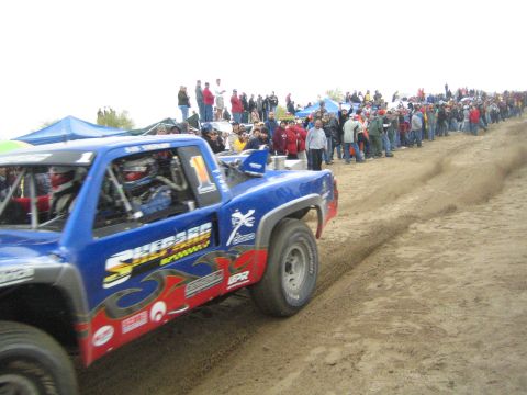 1990 Ford Bronco - blue off-road race truck numbered 1 with flame graphics drives along a sandy course past a long line of spectators.