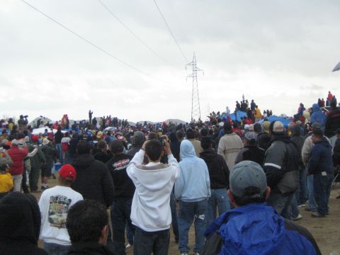 1990 Ford Bronco - Spectators in jackets and hoodies watch toward a power-line tower across a broad sandy clearing on a gray day.