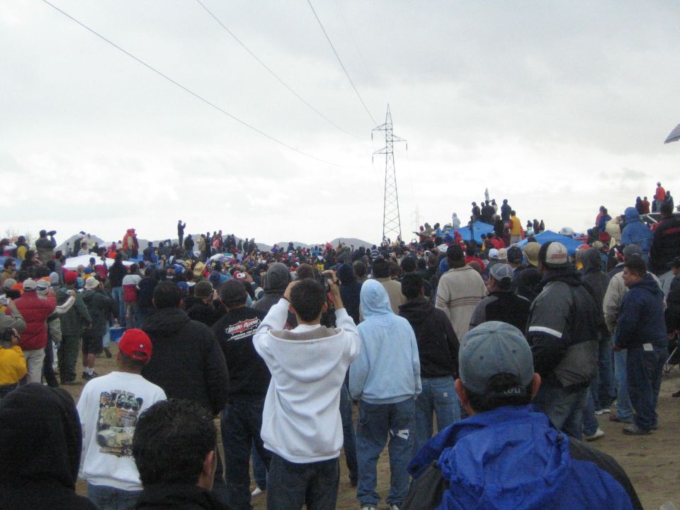 Crowd watching toward a power-line tower across a sandy expanse