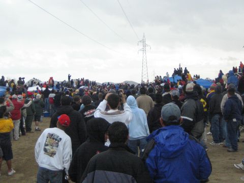 1990 Ford Bronco - dense crowd of bundled spectators faces a distant power-line tower across a sandy field scattered with tents.