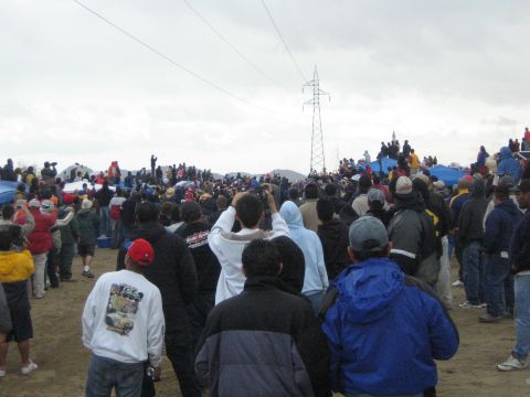 1990 Ford Bronco - Spectators in winter clothing and caps watch across a sandy desert field toward a power-line tower under gray clouds.