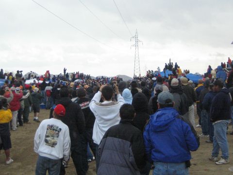 1990 Ford Bronco - crowd of jacketed spectators gathers on sandy ground watching toward a power-line tower beneath an overcast sky.