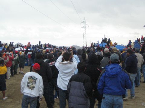 1990 Ford Bronco - Spectators in hoodies and jackets watch toward a power-line tower across sandy terrain dotted with pop-up tents.