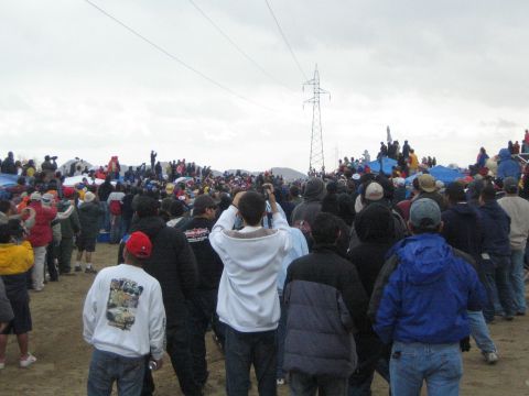 1990 Ford Bronco - crowd of spectators in caps and jackets faces a power-line tower across a sandy desert clearing on an overcast day.