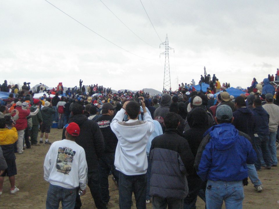 Crowd facing a power-line tower across a sandy desert clearing