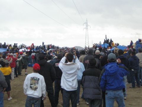 1990 Ford Bronco - large spectator crowd stands on sandy ground watching toward a power-line tower beneath a heavy gray sky.