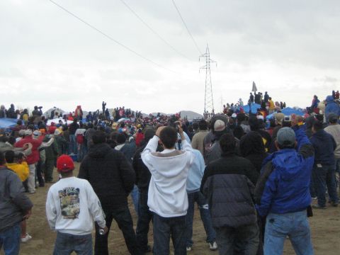 1990 Ford Bronco - Spectators photograph and watch across a sandy field toward a power-line tower under a pale overcast sky.