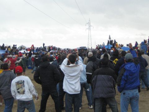 1990 Ford Bronco - crowd of bundled spectators watches across a sandy clearing where a white vehicle sits near a power-line tower.