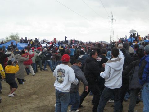 1990 Ford Bronco - dense crowd of spectators in jackets and caps gathers on a sandy desert course beneath power lines on an overcast day.