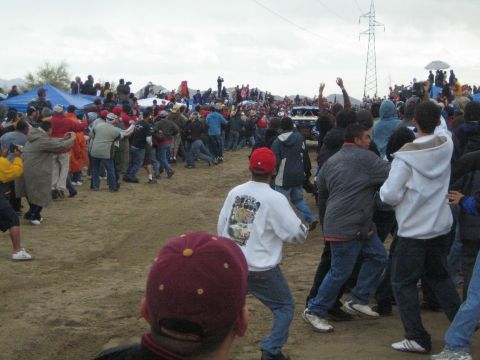 1990 Ford Bronco - crowd of fans presses toward the edge of a dirt course as a distant off-road race vehicle approaches under a gray sky.