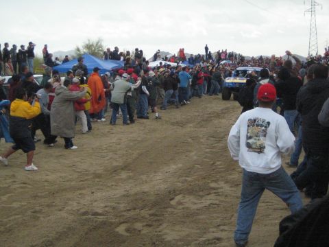 1990 Ford Bronco - Spectators in jackets and ponchos line a sandy desert course while a blue and yellow off-road truck passes in the background.