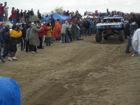 1990 Ford Bronco - blue and yellow off-road trophy truck rolls down a sandy desert course as crowds of fans watch from the sidelines.