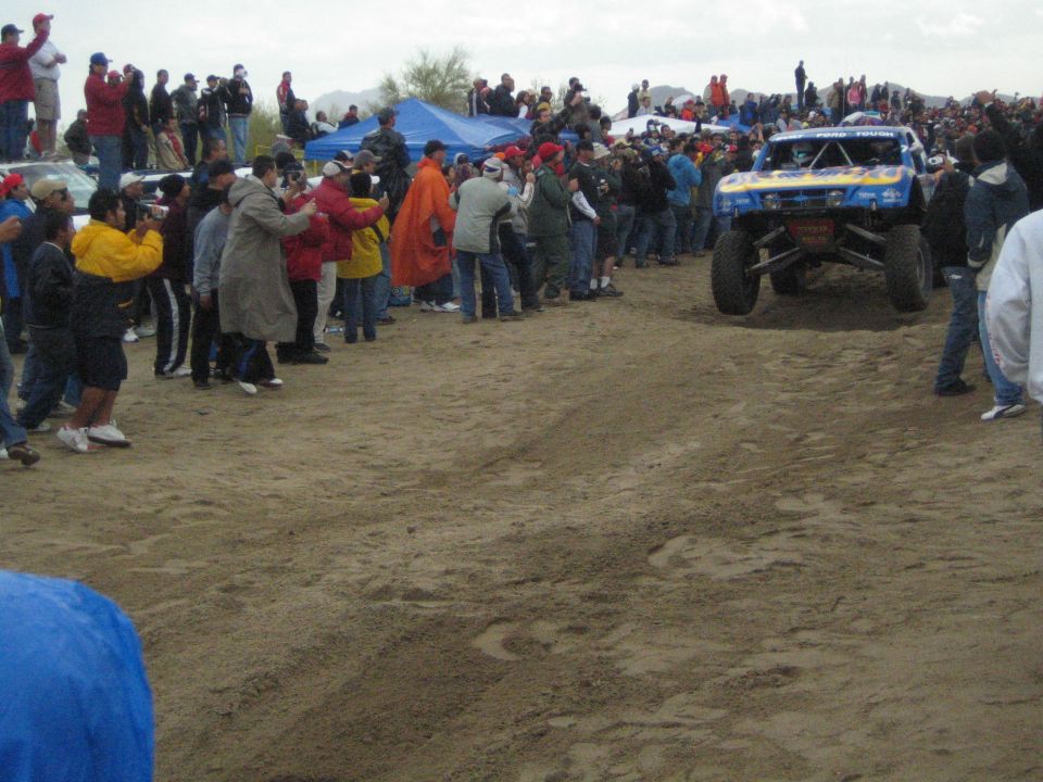Blue and yellow off-road trophy truck driving down a sandy course past crowds