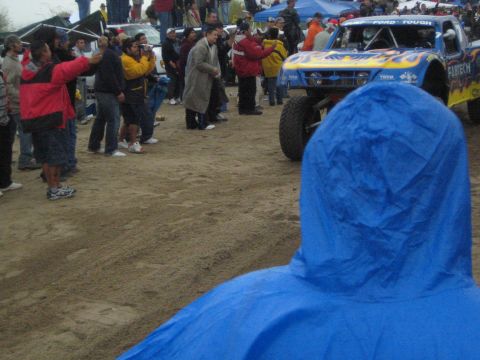 1990 Ford Bronco - blue off-road trophy truck with flame graphics and FORD TOUGH lettering sits among spectators, viewed past a person in a blue poncho.