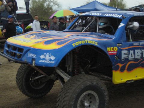 1990 Ford Bronco - blue off-road trophy truck with orange flames sports Toyo Tires, Fabtech and FORD TOUGH decals, with a driver in the cage and crowds behind.