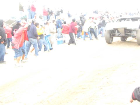 1990 Ford Bronco - bright, washed-out shot shows a white off-road truck passing a crowd of bundled-up spectators along a sandy course.