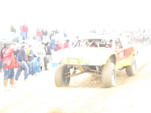 1990 Ford Bronco - washed-out action shot shows a red and yellow tube-frame off-road buggy on a sandy course as spectators watch from the left.