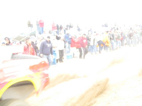 1990 Ford Bronco - heavily overexposed, motion-blurred shot shows a red and yellow race vehicle streaking past a long line of spectators on a dirt course.