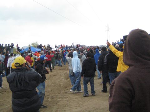 1990 Ford Bronco - Spectators in hoodies and jackets stand and walk along a sandy desert course, one raising a phone, beneath power lines and a gray sky.
