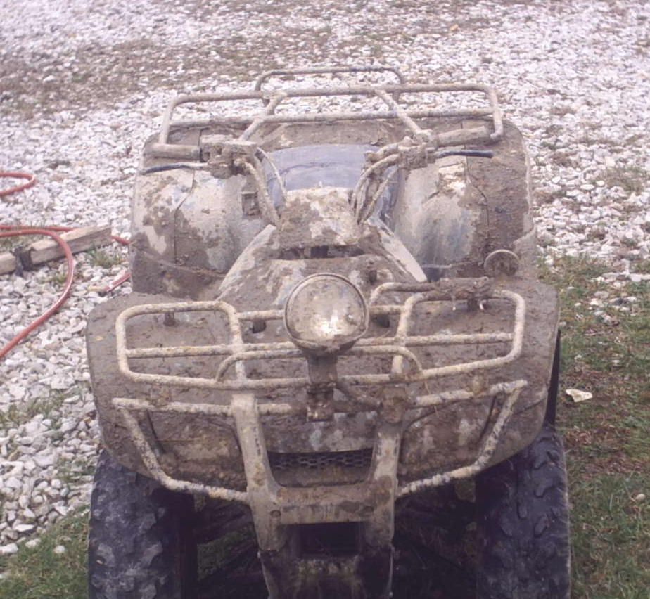 Muddy ATV parked on gravel. Front view shows handlebars, racks, and a headlight