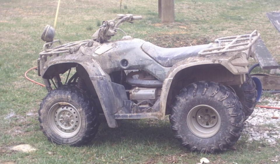 Mud-covered utility ATV parked side-on in a grassy yard with a hose lying nearby