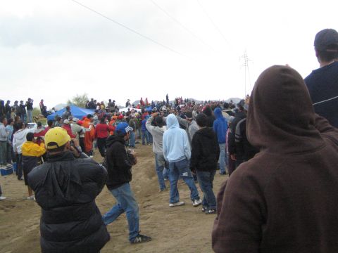 1990 Ford Bronco - crowd of bundled-up spectators lines a sandy desert course, viewed over the shoulder of a person in a brown hooded sweatshirt.