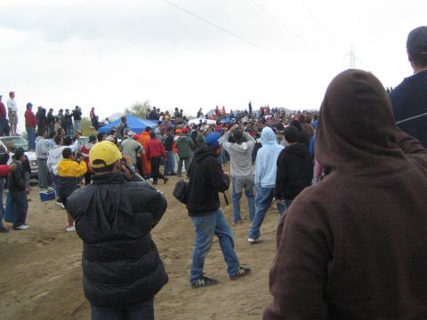 1990 Ford Bronco - Spectators in winter jackets and caps gather and walk on a sandy desert course, viewed past a person in a brown hoodie.