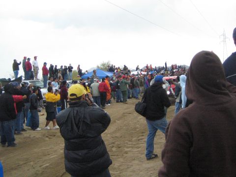 1990 Ford Bronco - Spectators stand on parked cars and crowd a sandy desert course beneath power lines and a gray overcast sky.
