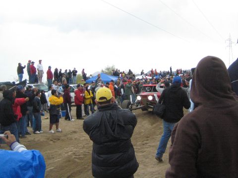 1990 Ford Bronco - red tube-frame off-road buggy with its headlights on idles among a crowd of bundled spectators on a sandy desert course.