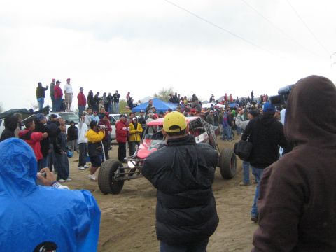 1990 Ford Bronco - red tube-frame off-road buggy sits surrounded by a crowd of spectators on a sandy desert course under a gray sky.