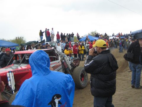 1990 Ford Bronco - red number 4 tube-frame off-road buggy with a BFGoodrich decal sits among spectators, one in a blue orca-print poncho in front.