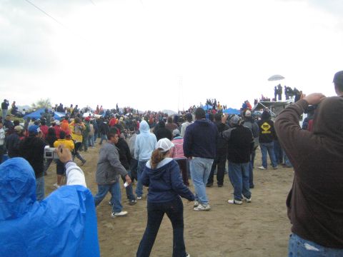 1990 Ford Bronco - crowd of bundled spectators gathers on a sandy desert course as one in the foreground raises a phone to take a photo.
