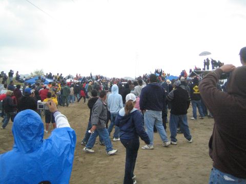 1990 Ford Bronco - crowd of spectators walks across a sandy desert course toward a larger gathering, with people perched on a vehicle in the distance.