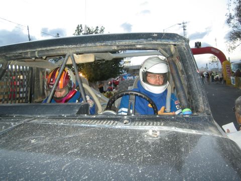 1990 Ford Bronco - Two helmeted occupants sit inside the roll-caged cab of a dust-covered off-road race vehicle near a banner-marked staging arch.