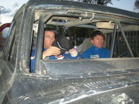 1990 Ford Bronco - Two co-drivers in blue racing suits sit inside the dusty roll-caged cab of an off-road race truck.