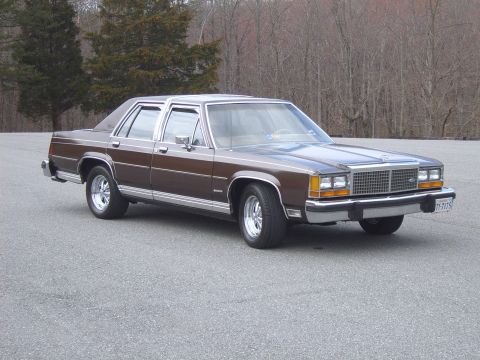 1982 Ford Crown Victoria LTD - This sedan features a prominent chrome grille and distinctive body lines, sitting on a paved surface with trees in the background.